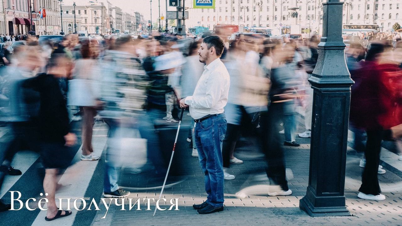 Презедент фонда Максим стоит в центре кадра боком с тростью. Мимо него проходит толпа людей с эффектом размытия в движении. У ног надпись «Всё получится»